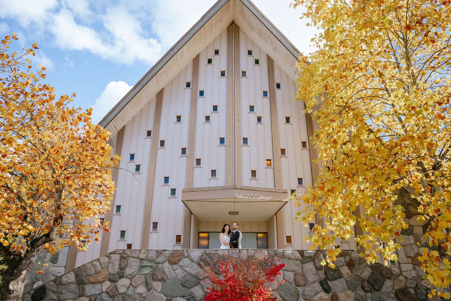 Couple standing in the entrance of a tall white chapel with vertical wood accents, framed by golden autumn trees and a stone wall.