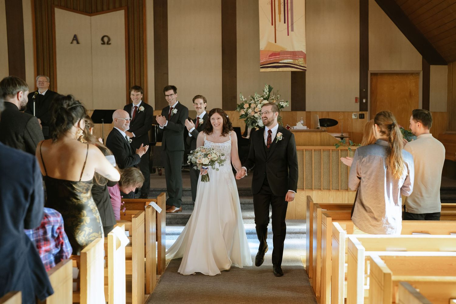 Michael and Melissa walking hand in hand down the church aisle as newlyweds, past smiling guests in wooden pews.