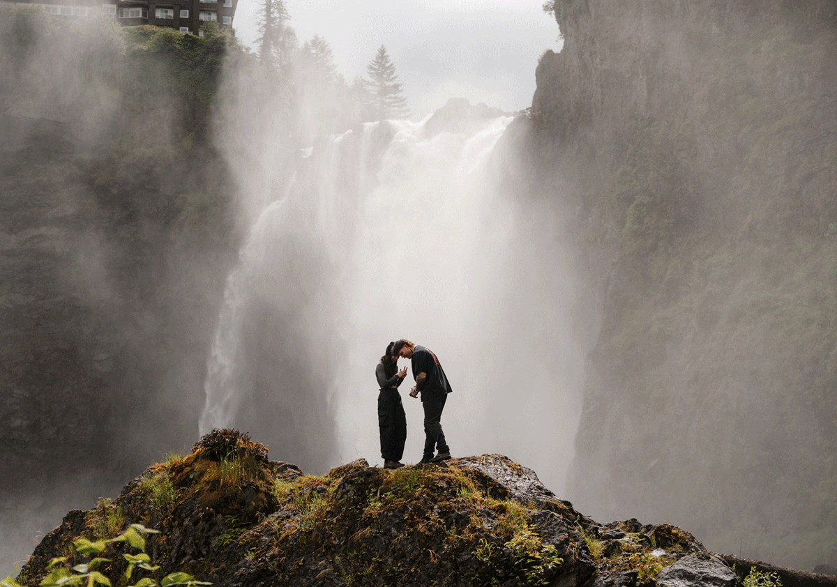 Couple sharing a romantic moment on a mossy rock ledge in front of a majestic misty waterfall.