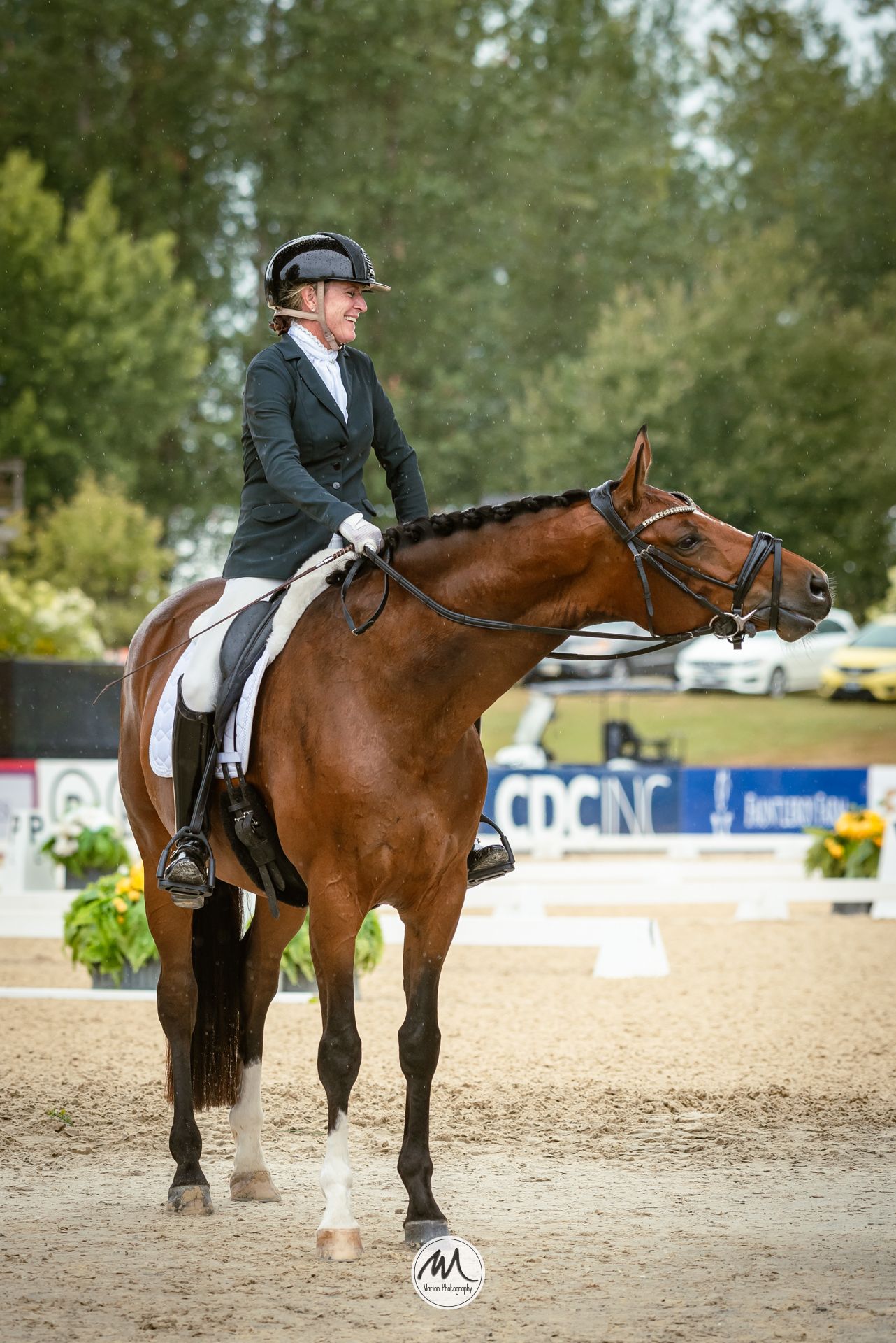 A female equestrian in a black helmet and green jacket rides a bay horse in a dressage competition on a sandy arena.
