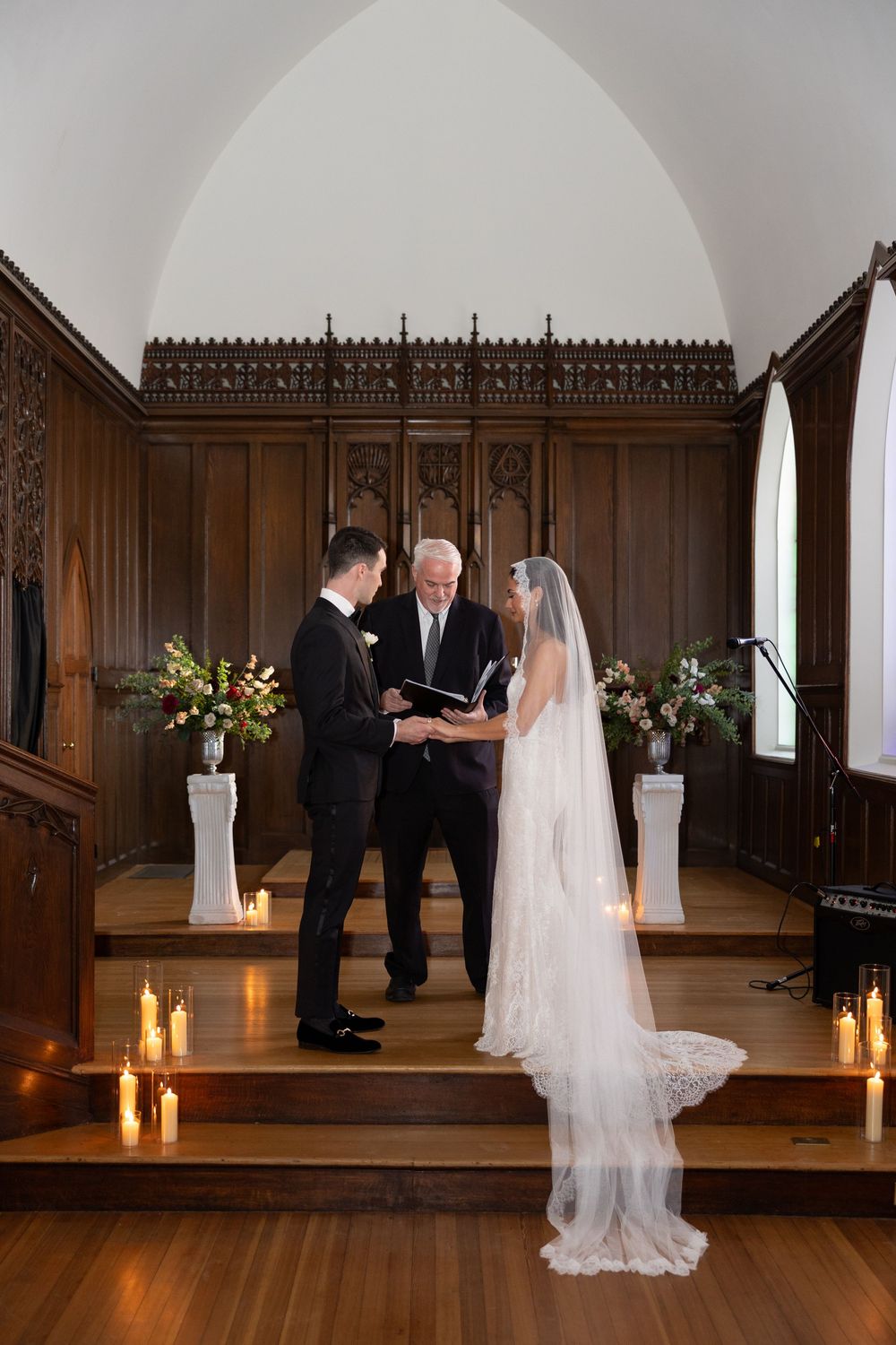 Bride and groom exchange vows at candlelit chapel altar with officiant, wooden panels, and floral arrangements.