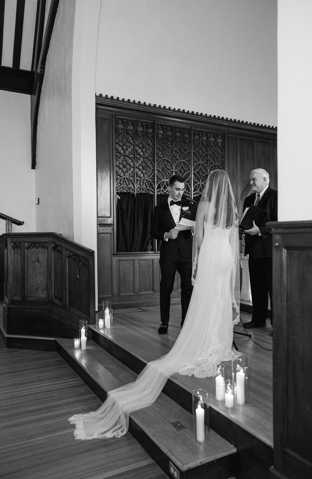 Black and white wedding ceremony photo of bride in flowing veil facing groom in tuxedo, with candles lining the altar steps.