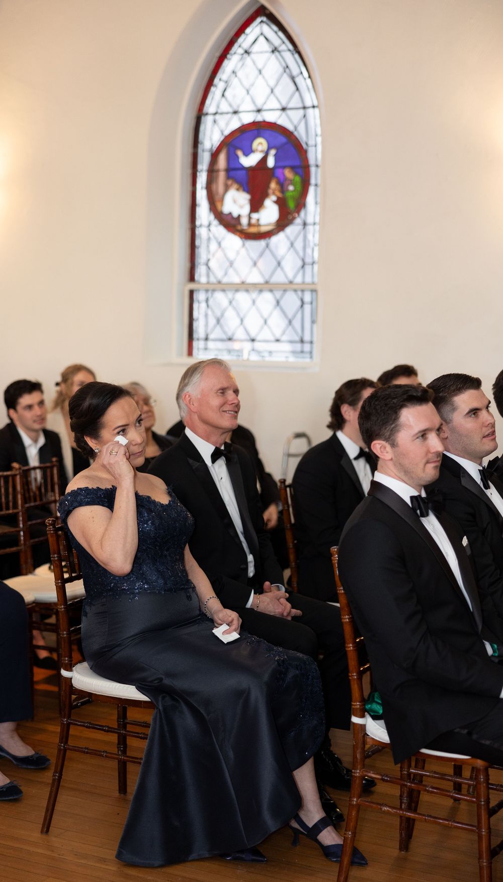Guests in formal attire seated in a chapel during a wedding ceremony, with a stained glass window behind them.