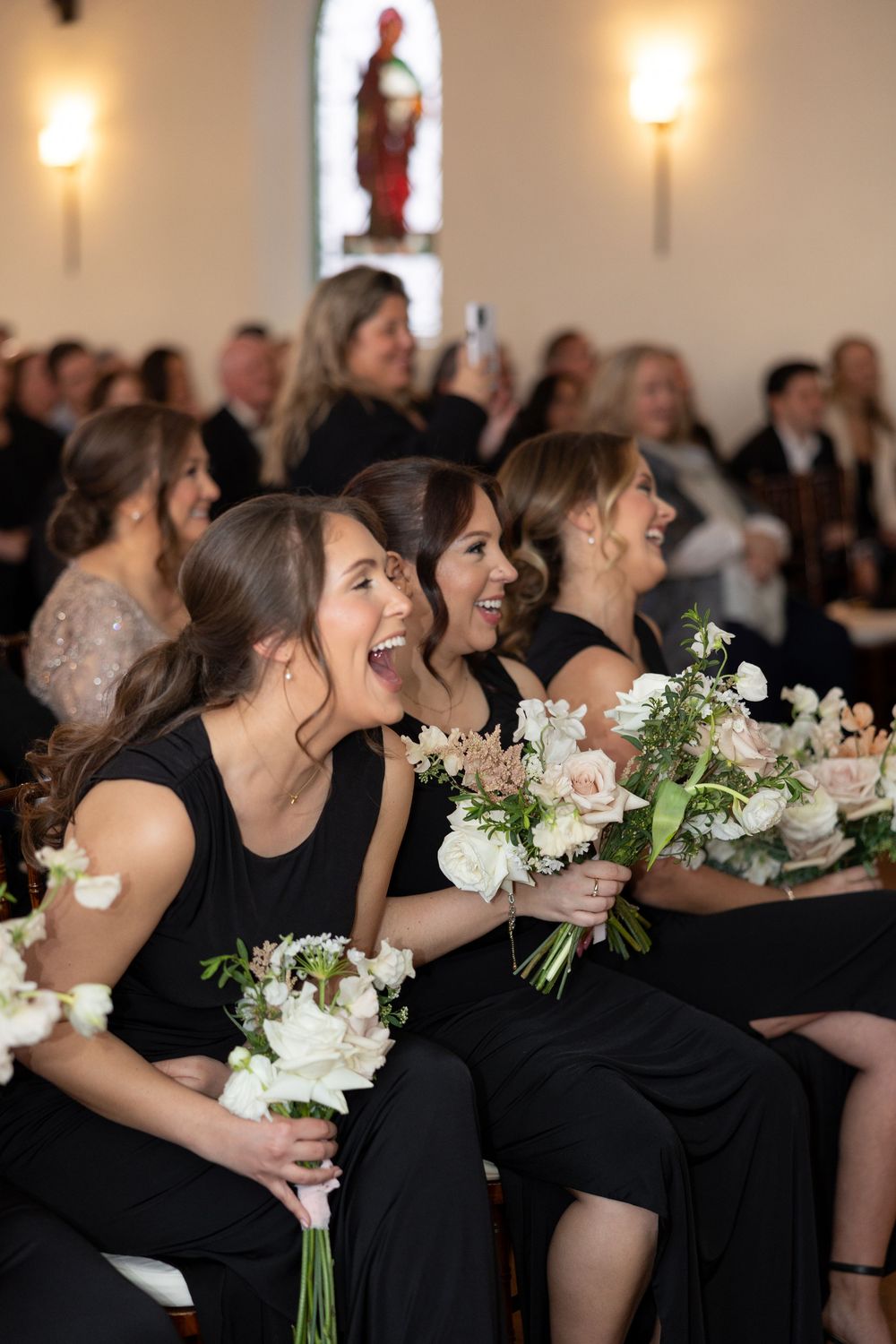 Bridesmaids in black dresses laughing and holding white floral bouquets during a wedding ceremony in a church.