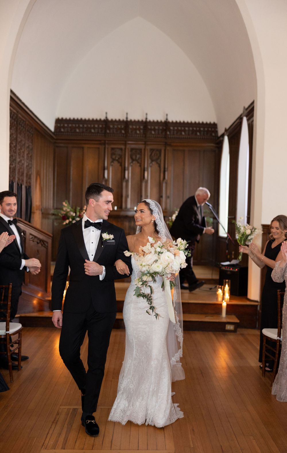 Bride and groom walk down the aisle of an elegant chapel after their wedding ceremony, smiling joyfully.