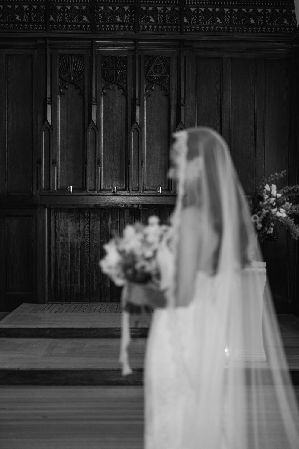 Black and white photo of a bride in a veil holding a bouquet standing before ornate wooden church altar panels.