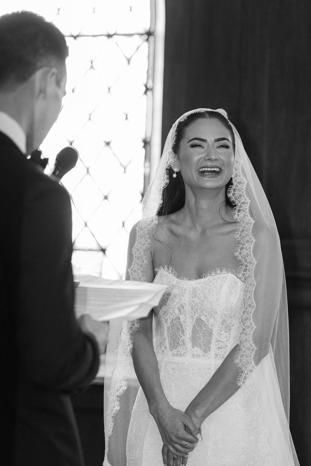 Black and white photo of a radiant bride laughing joyfully during wedding ceremony, wearing lace veil and strapless gown.