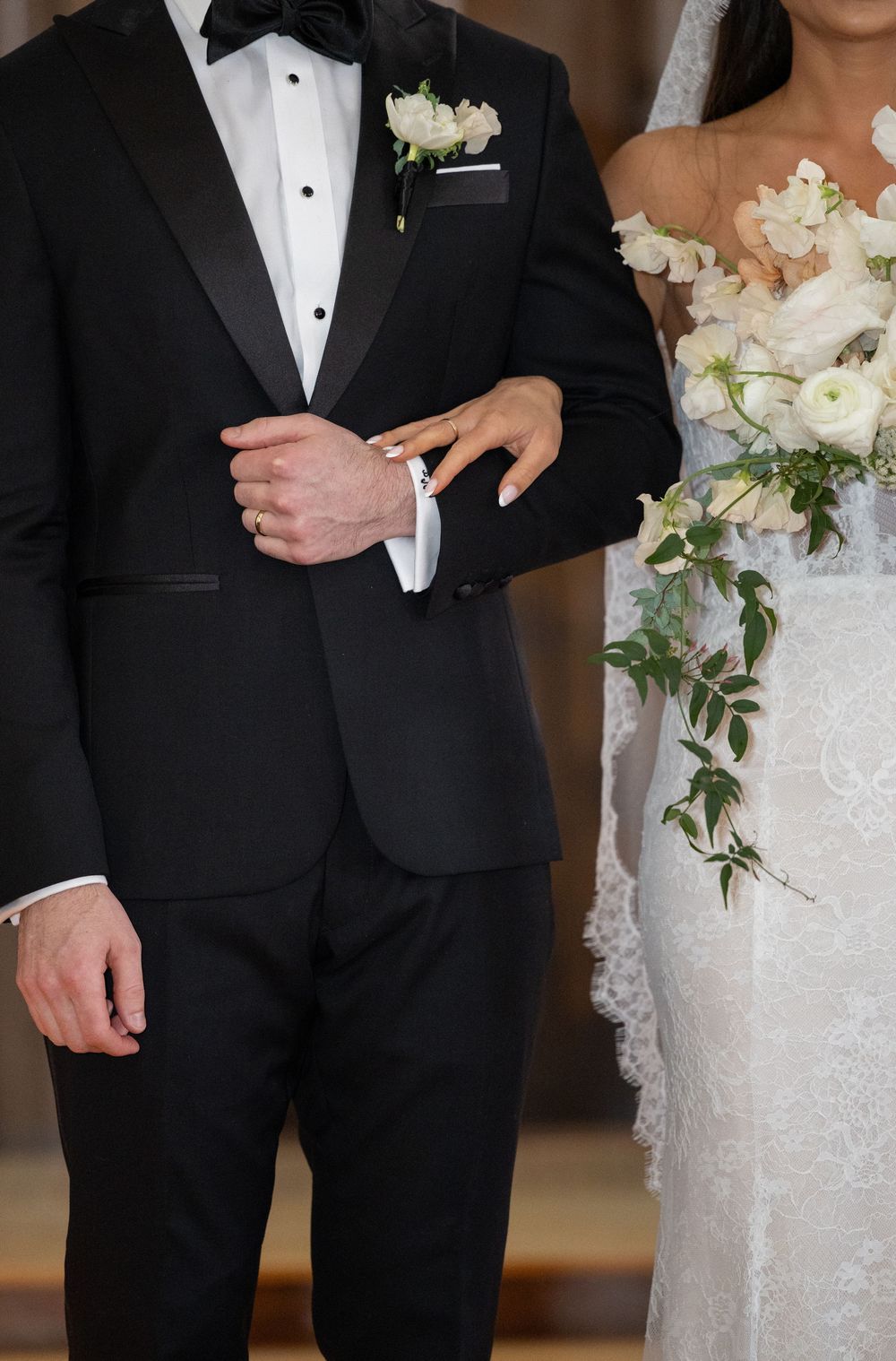 Bride in lace gown holding white floral bouquet stands beside groom in black tuxedo with boutonniere.