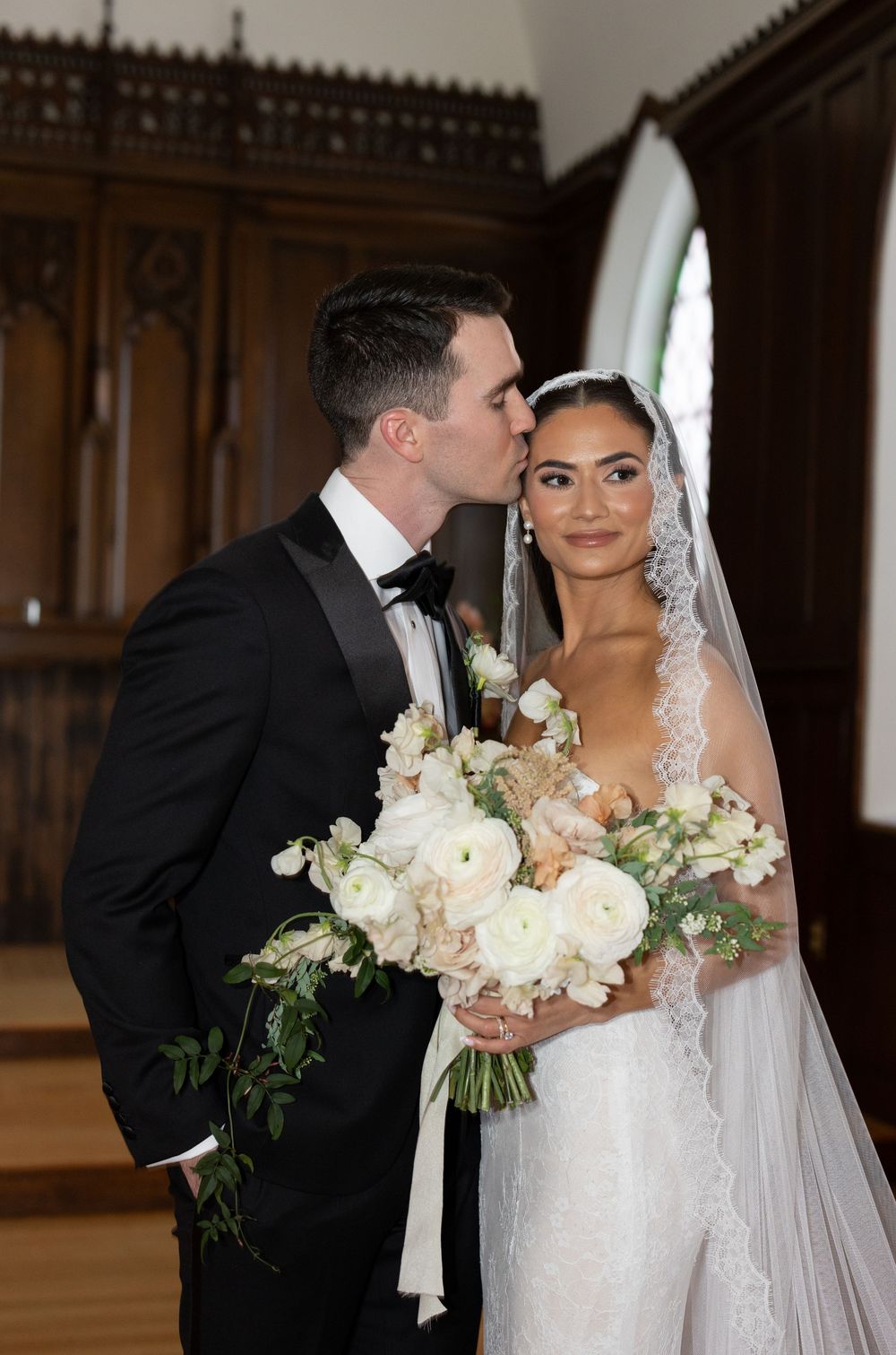 Groom in black tuxedo kisses bride wearing lace wedding dress and veil holding white floral bouquet in elegant church.