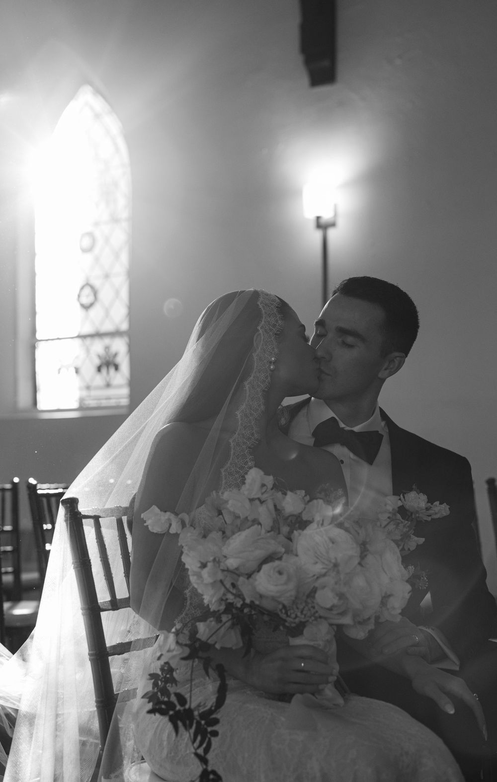 Black and white wedding photo of bride and groom sharing a kiss in a church, bride holding a bouquet.