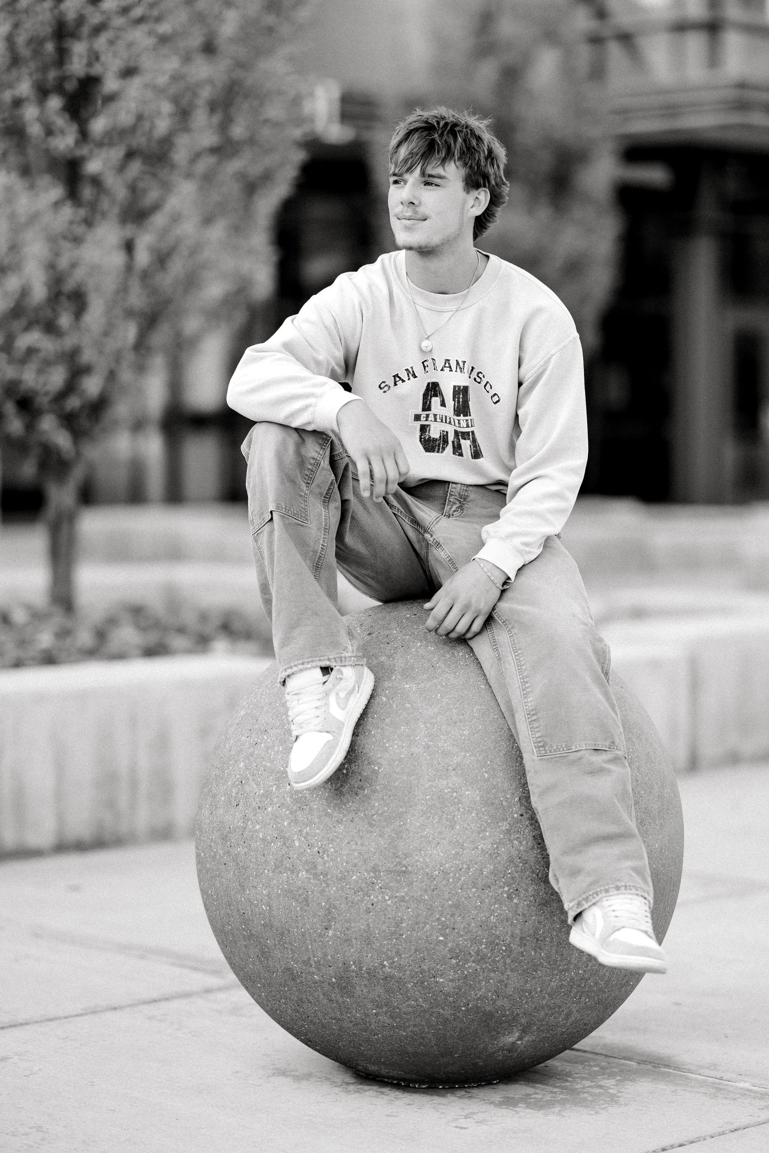 Young man in San Francisco sweatshirt and jeans sitting on a large stone sphere in an urban plaza, black and white photo.