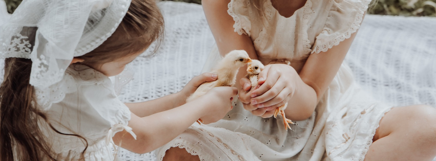 Two girls in white lace dresses gently hold a baby chick together outdoors.