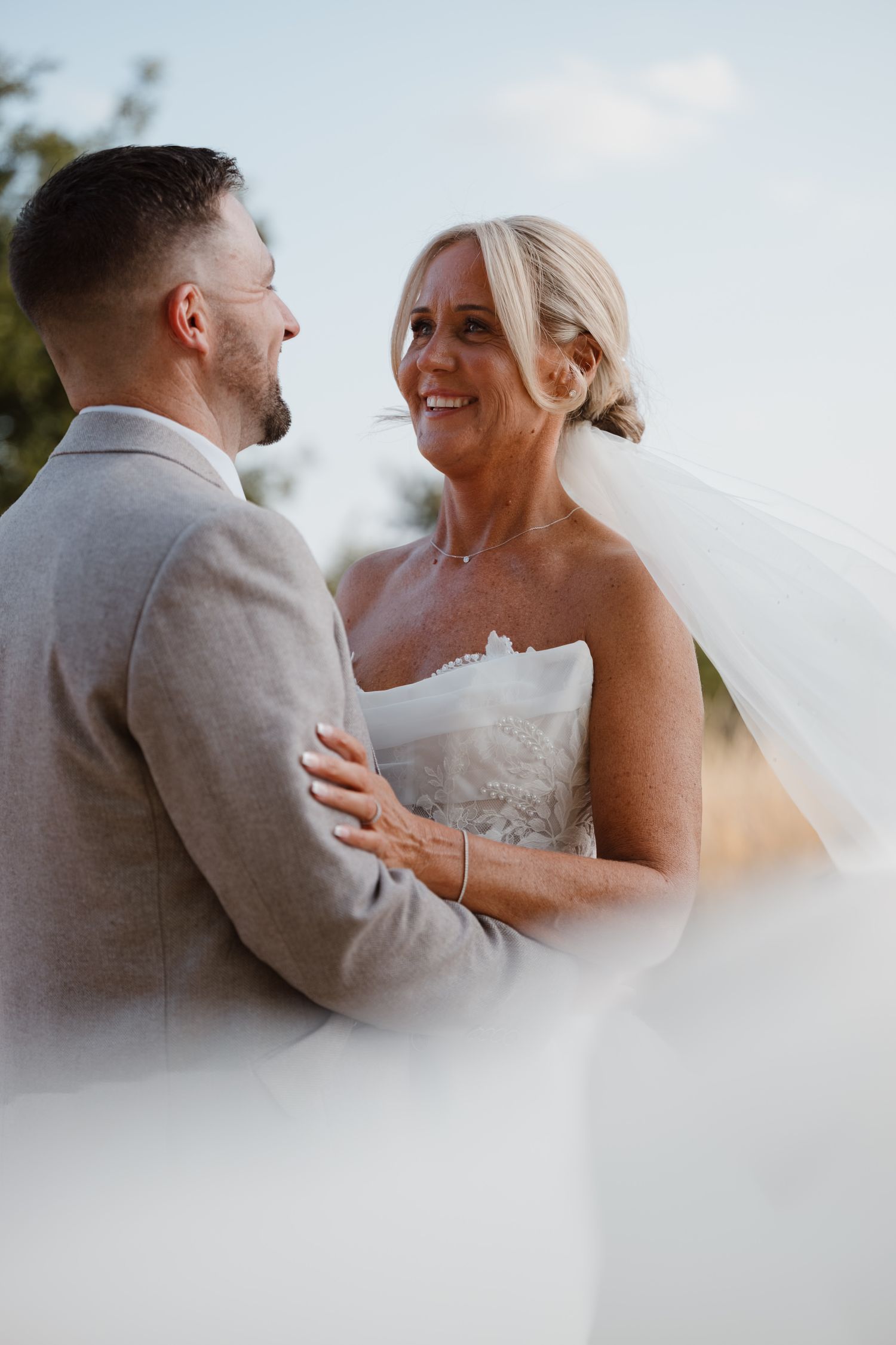 Bride and groom share a romantic moment outdoors, her veil flowing, as she smiles in a strapless gown and he wears a gray suit.