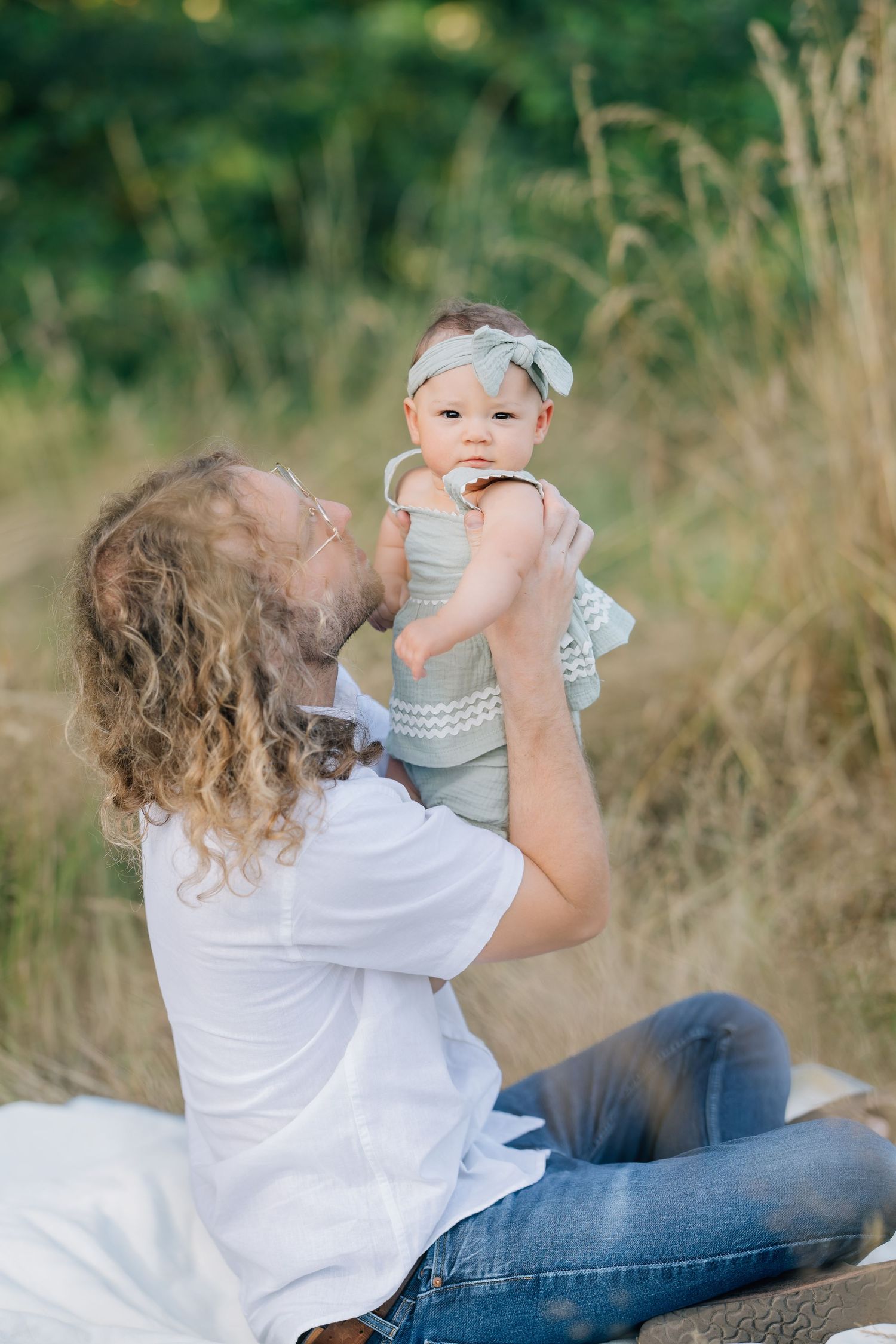 A father with curly hair lifts his baby girl outdoors in a sunny field, sharing a joyful moment together.