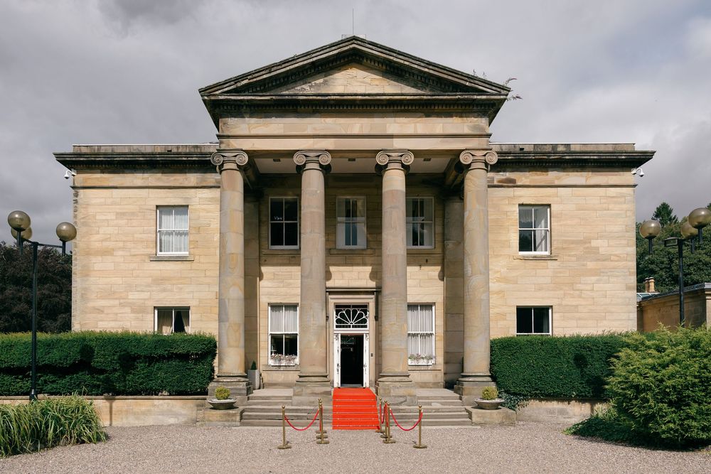 Grand neoclassical stone building with Greek columns, red carpet entrance, and manicured hedges under cloudy skies.