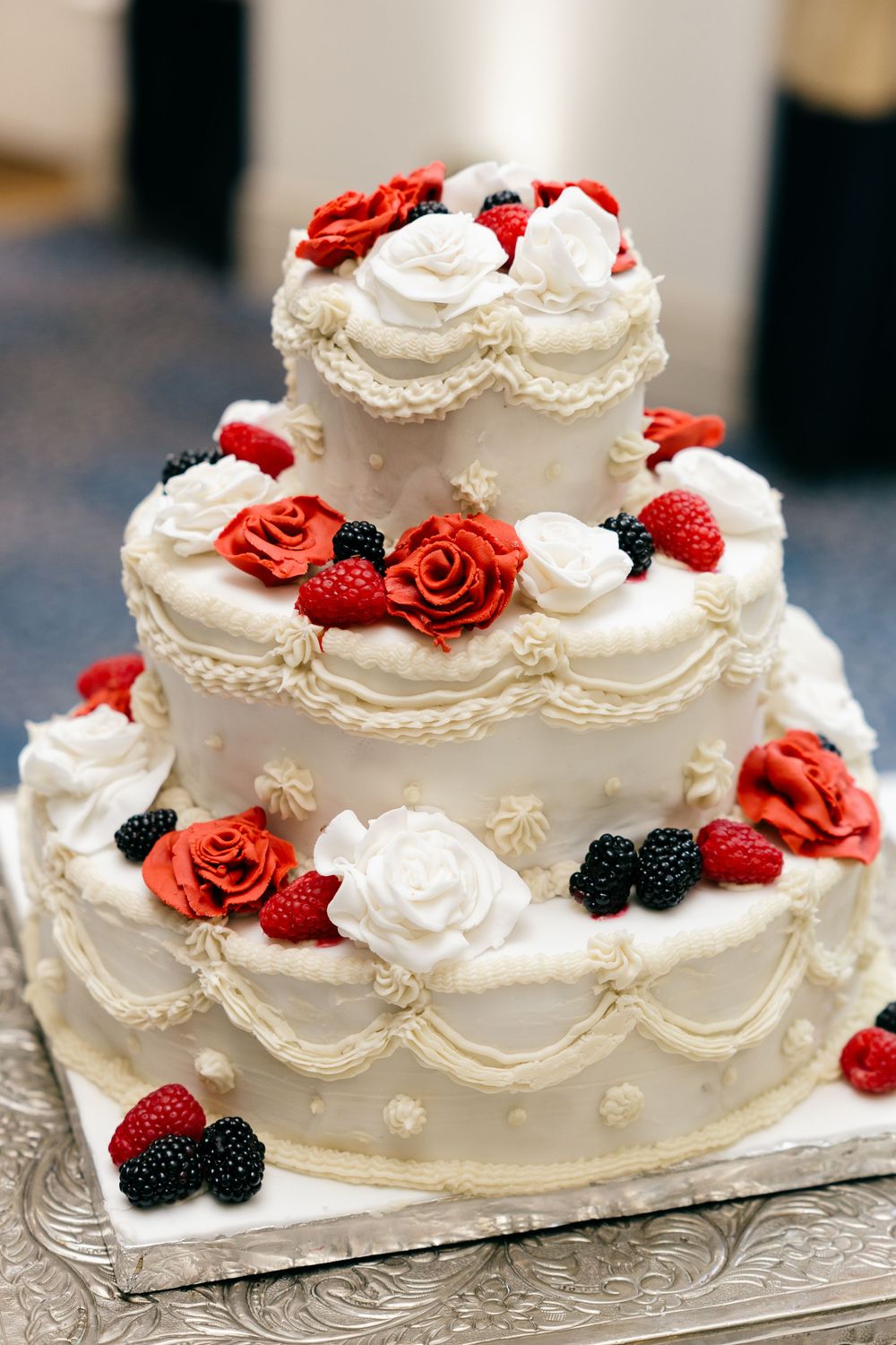 Three-tier white wedding cake decorated with red roses, raspberries, blackberries, and white frosting swags on a silver stand.