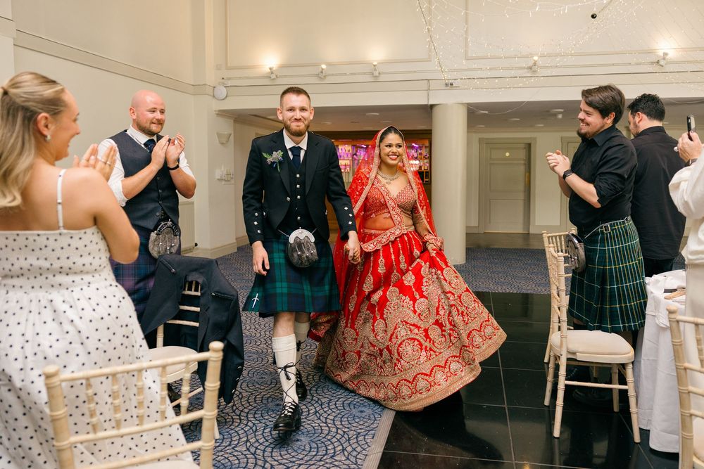 Bride in red lehenga and groom in Scottish kilt walk together at multicultural wedding ceremony with guests applauding.