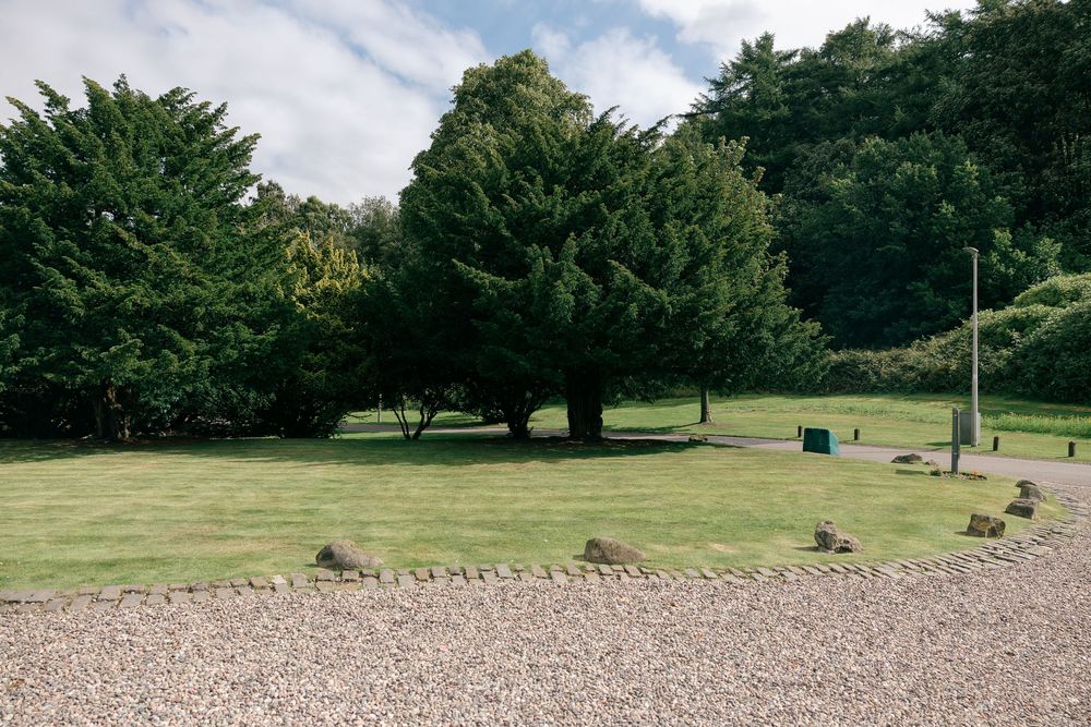Peaceful garden grounds with large green trees, manicured lawn, and gravel path on a sunny day.