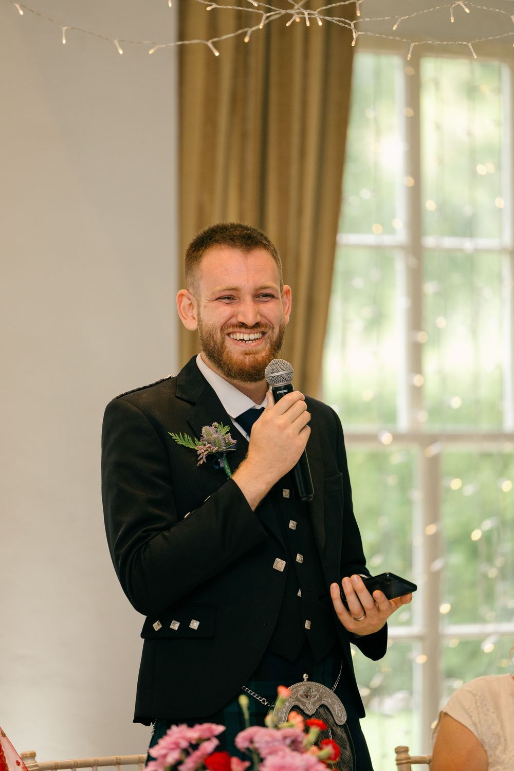 Smiling groom in Scottish Highland dress giving a wedding speech while holding a microphone and phone.