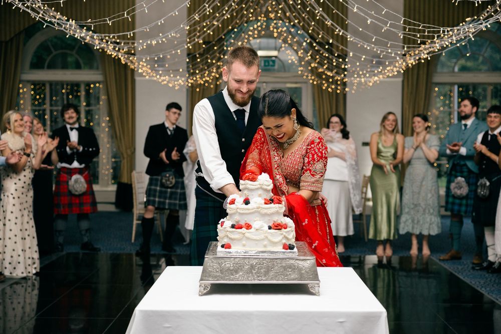 Bride in red sari and groom in vest cut wedding cake at elegant venue with fairy lights and guests in kilts.