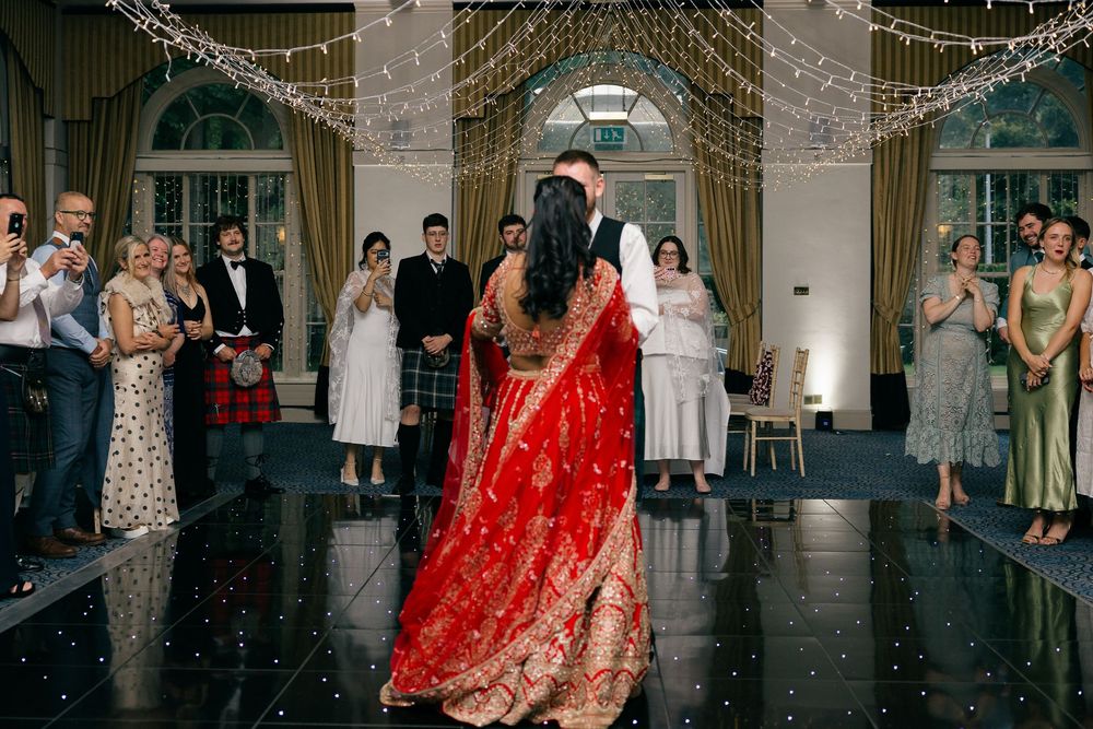 Bride in red lehenga and groom share first dance at elegant wedding reception with fairy lights and guests watching.