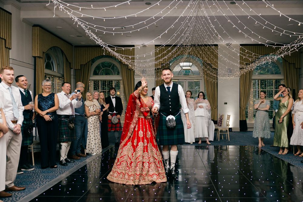Bride in red lehenga and groom in Scottish kilt share first dance at elegant wedding reception with fairy lights.