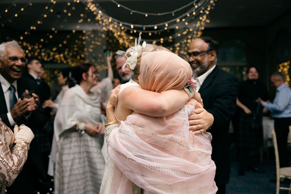 Two guests share a warm hug at a wedding reception with fairy lights and smiling attendees in the background.