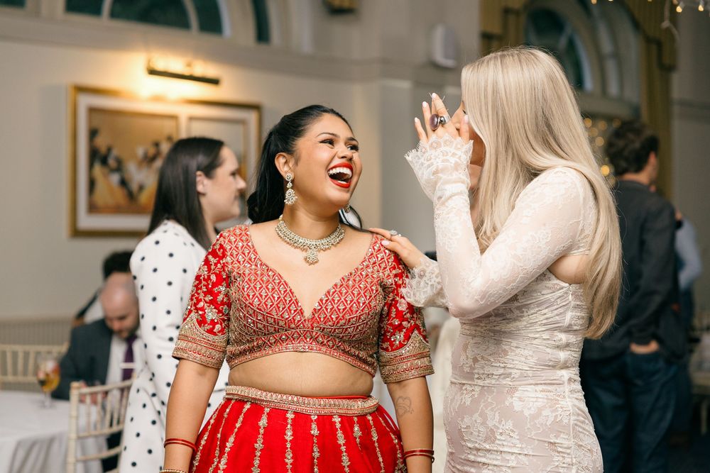 Two women laughing joyfully at an Indian wedding, one in a red embroidered lehenga and another in a white lace dress.