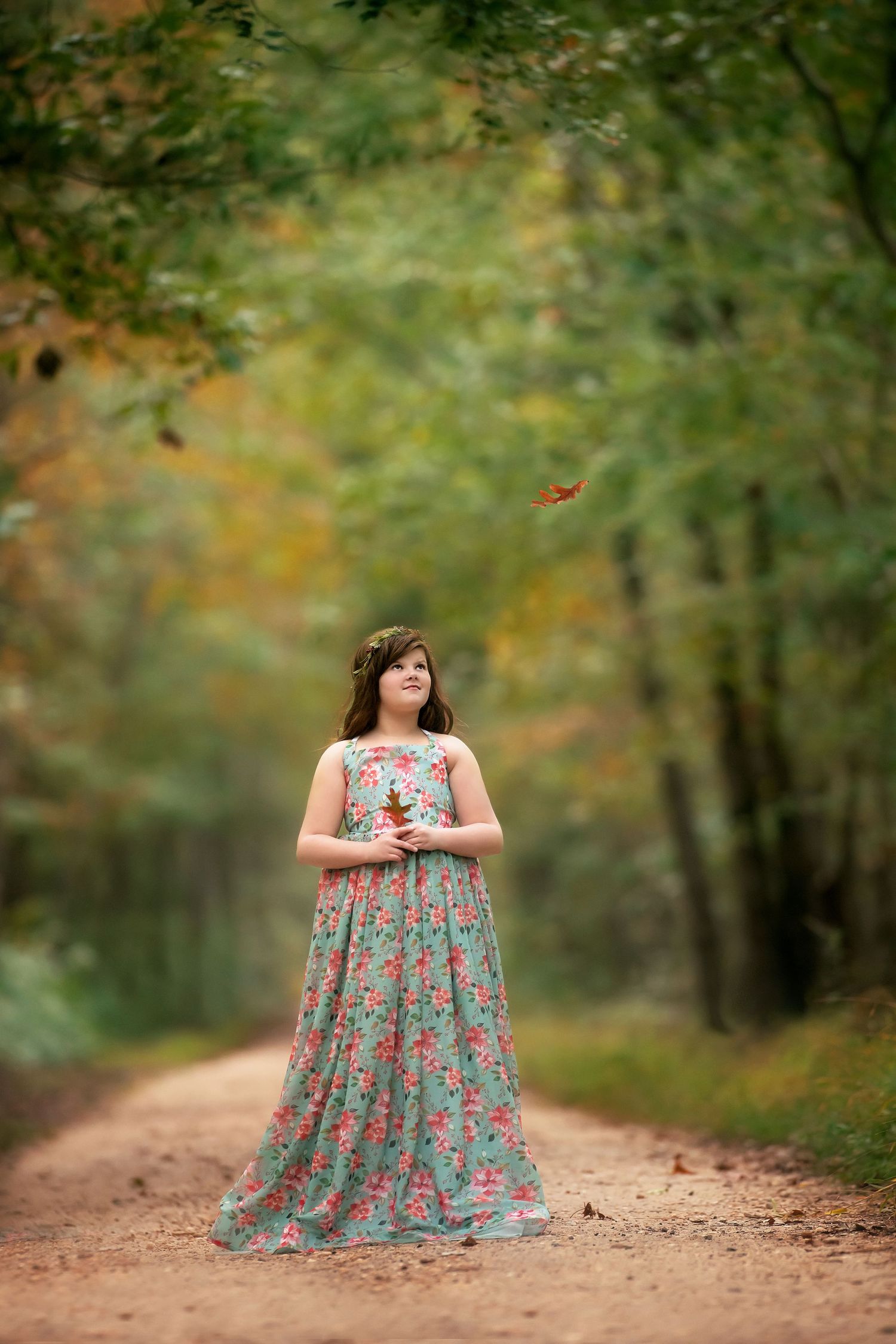 Girl in a floral maxi dress holding a leaf stands on a dirt path in an autumn forest as a leaf falls nearby.