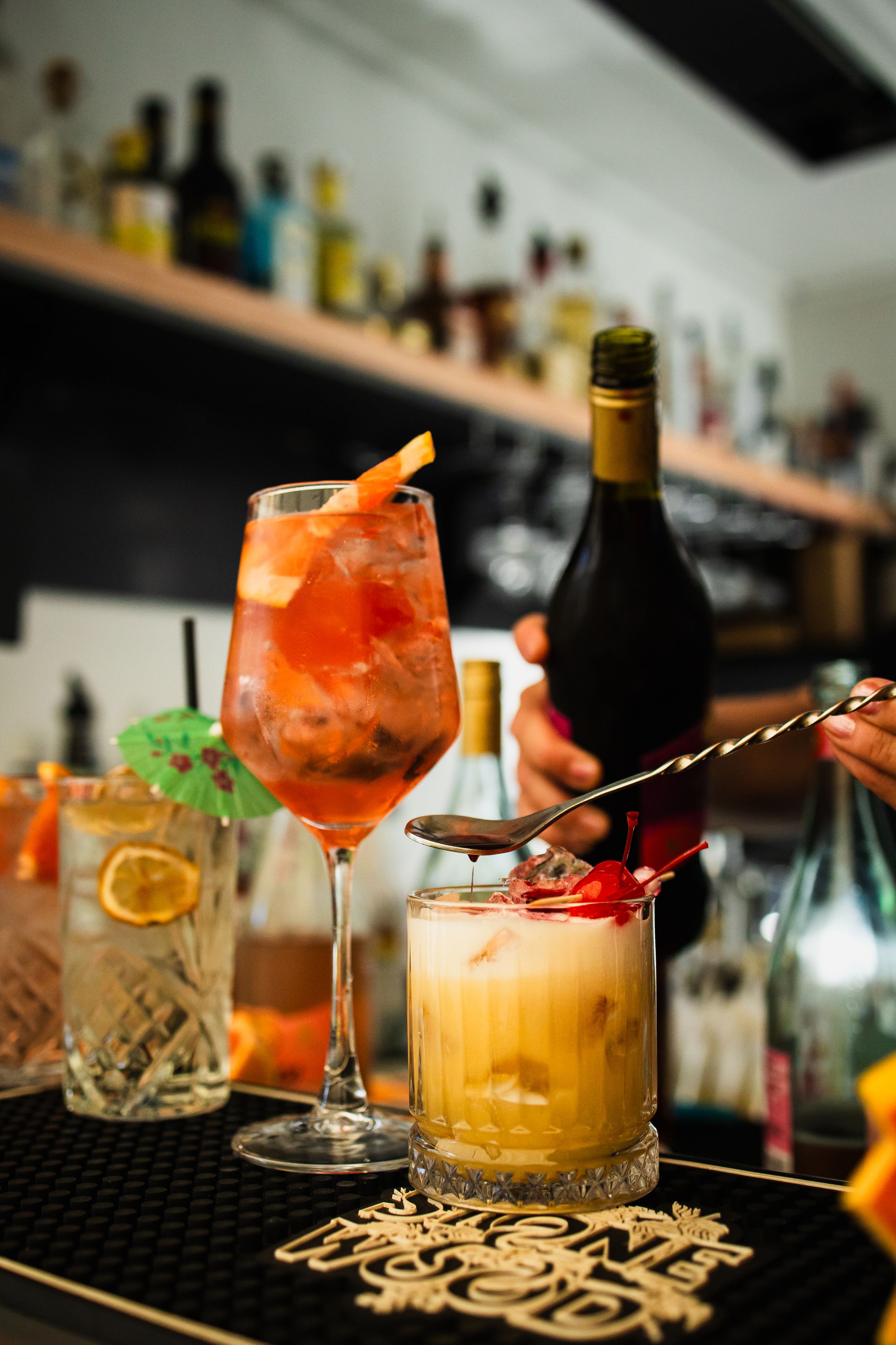 Colorful cocktails on a bar counter, including an Aperol Spritz in a wine glass with orange garnish and a creamy drink.