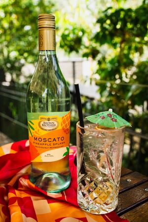Brown Brothers Moscato Pineapple Splash wine bottle beside a cocktail glass with umbrella on outdoor table.