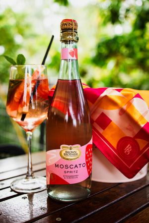 Brown Brothers Moscato Spritz Strawberry & Hibiscus bottle beside a wine glass and gift box outdoors.