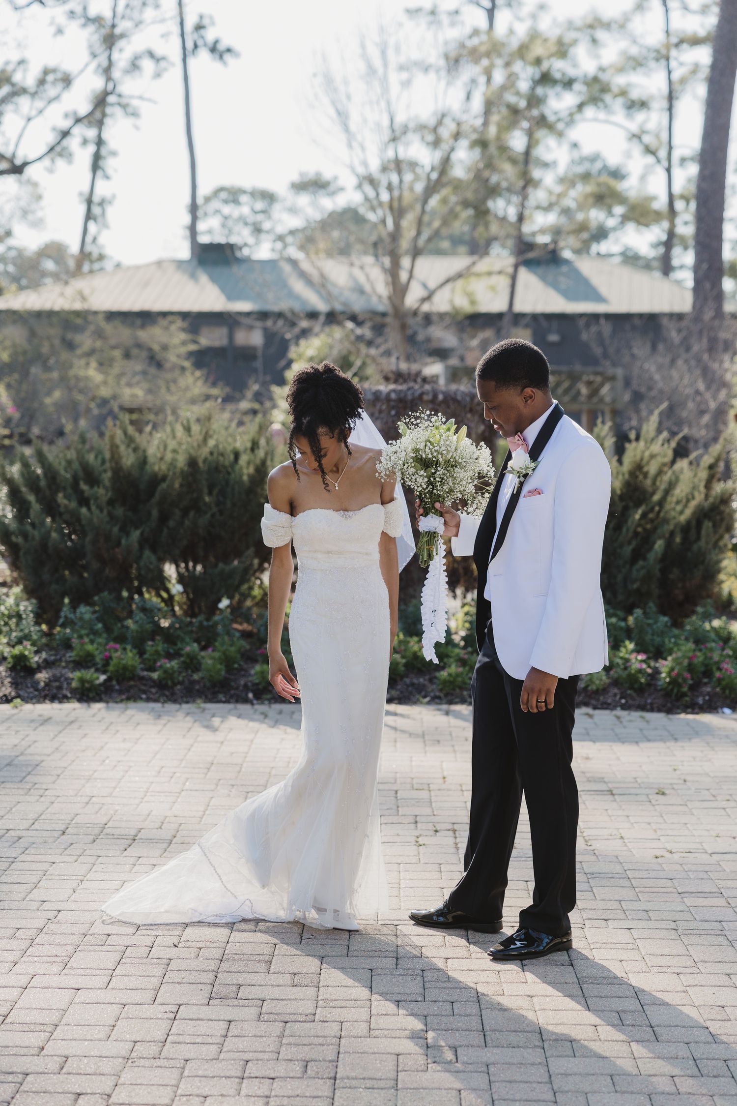 Bride in off-shoulder white gown and groom in white tuxedo jacket share a romantic moment on a brick patio.