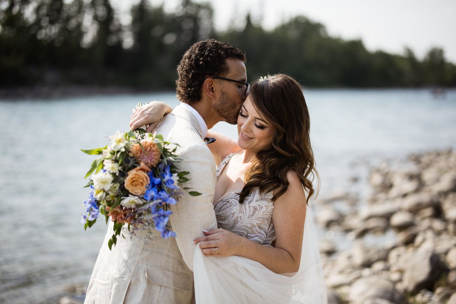 Groom kissing the bride's forehead by the river as she holds a blue and peach bouquet