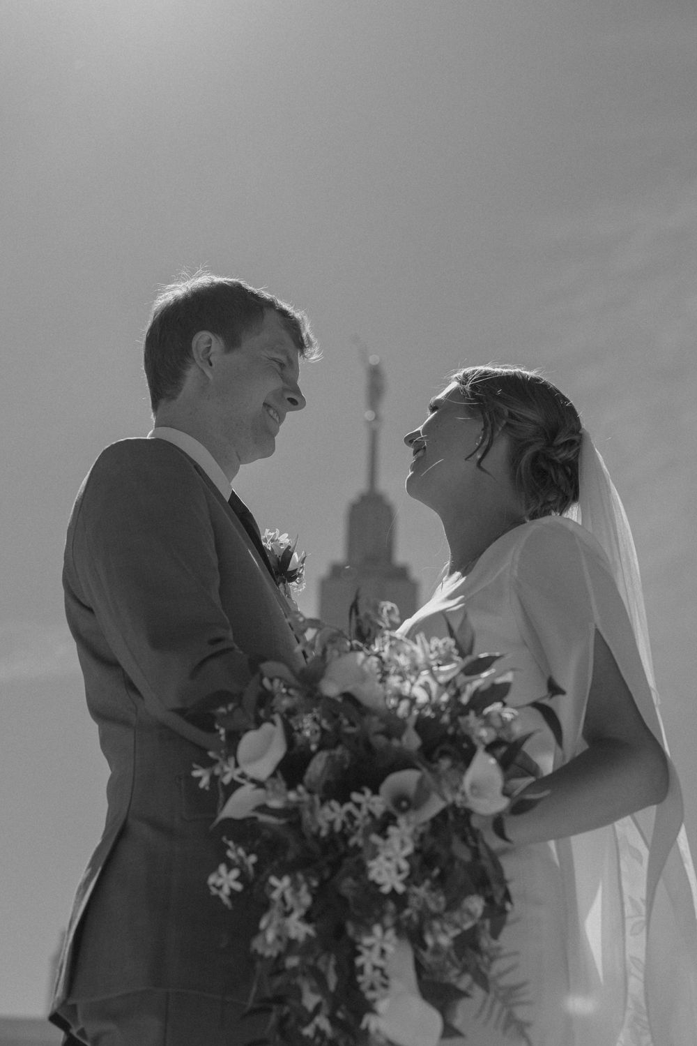 Black and white wedding photo of smiling bride and groom holding bouquet with church steeple in background.
