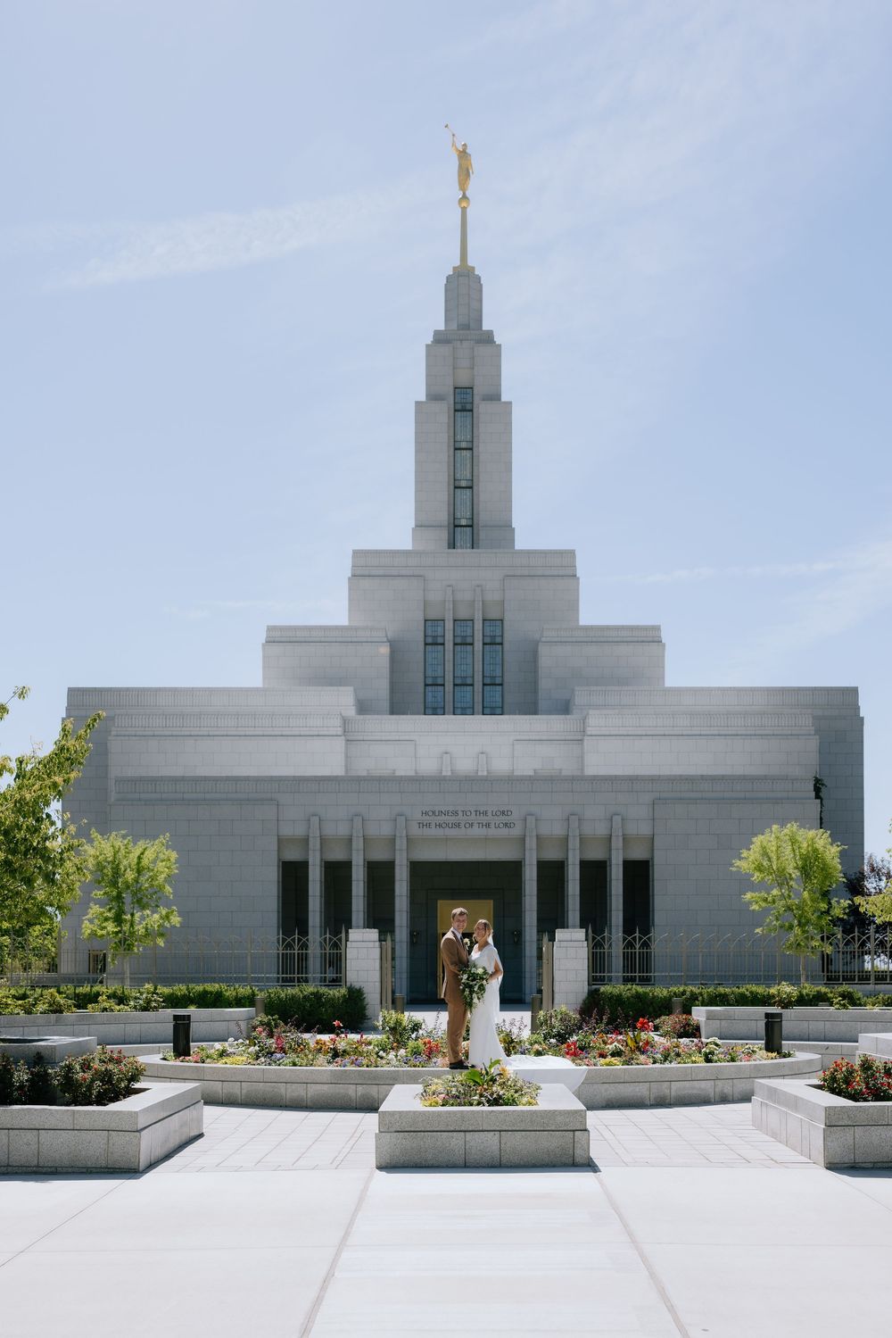 Bride and groom pose before a white LDS temple with golden spire and manicured gardens on a sunny day.