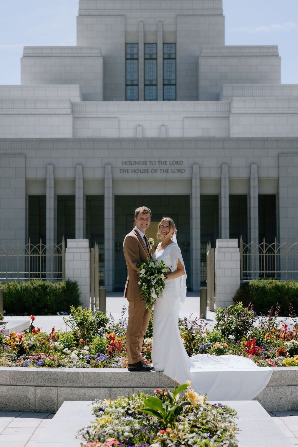 Bride and groom pose in front of an LDS temple with lush flower gardens on their wedding day.