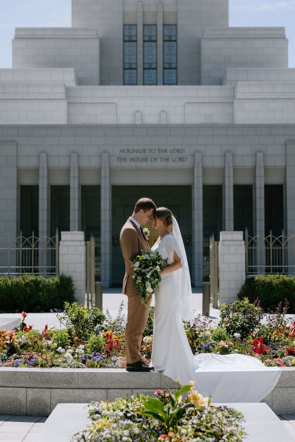 Bride and groom embrace in front of an LDS temple surrounded by colorful flower gardens on their wedding day.