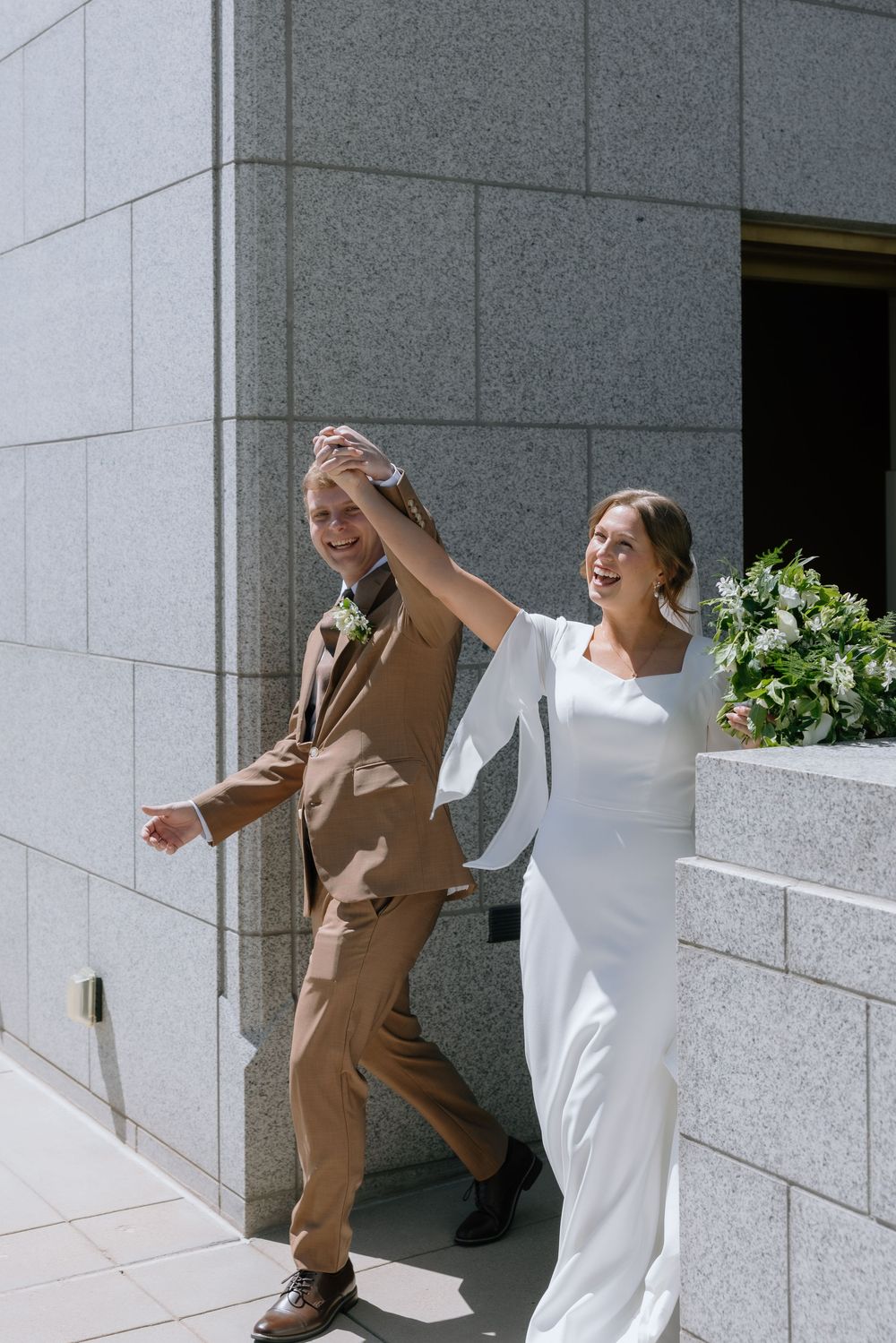 Joyful newlyweds exit a building, bride in white gown raising groom's hand, groom in brown suit, on a sunny day.
