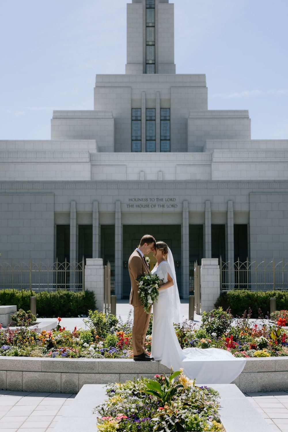 Bride and groom embrace in front of an LDS temple surrounded by colorful gardens on their wedding day.