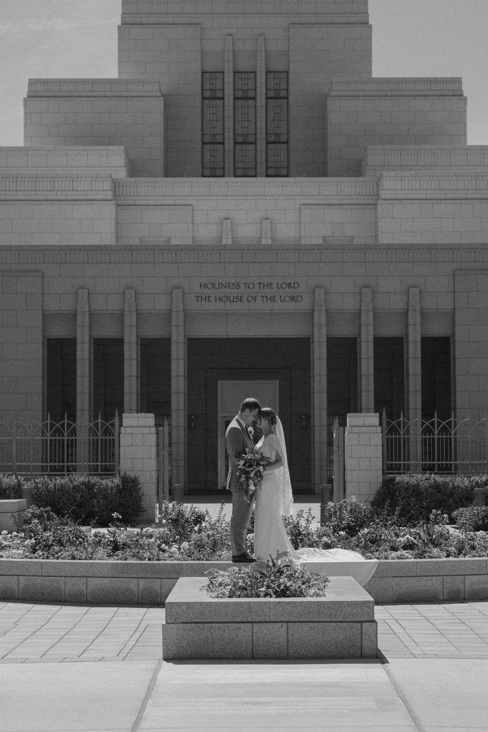 Black and white photo of bride and groom kissing in front of an LDS temple with lush garden.