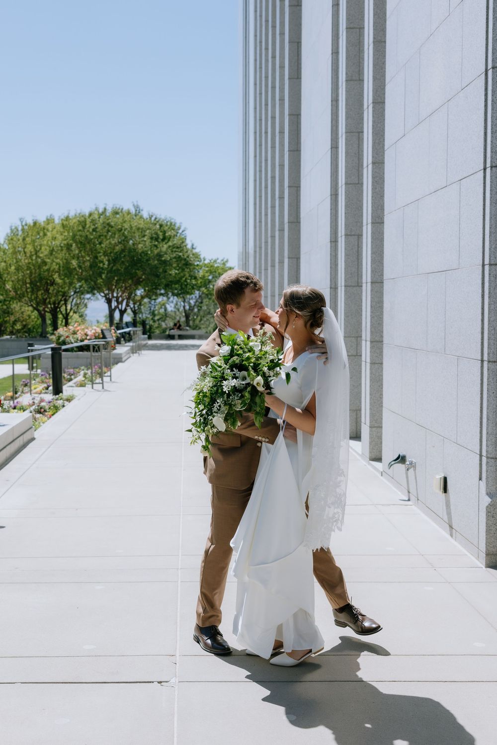 Bride and groom share a romantic moment outside a white stone temple, holding a lush green bouquet on their wedding day.