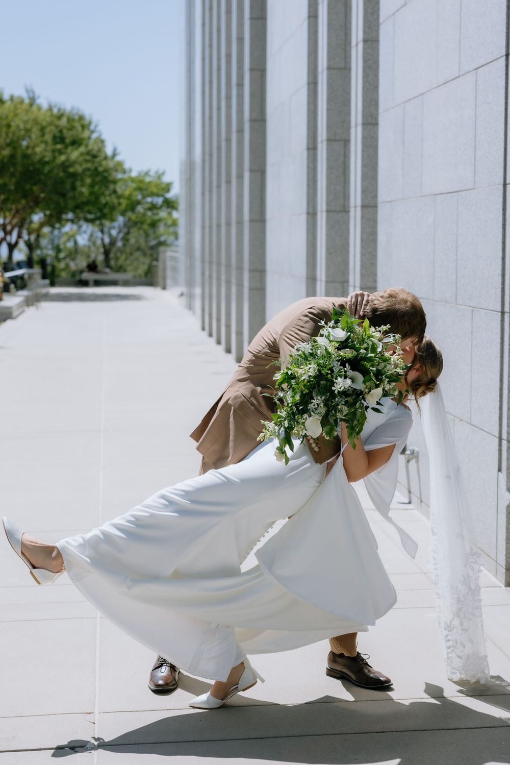 Bride in white dress and groom in tan suit share a romantic dip kiss outside a grand marble building.