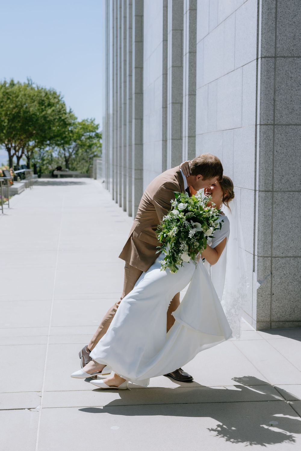 Bride and groom share a romantic dip kiss beside white marble columns, bride holding lush greenery bouquet in white gown.