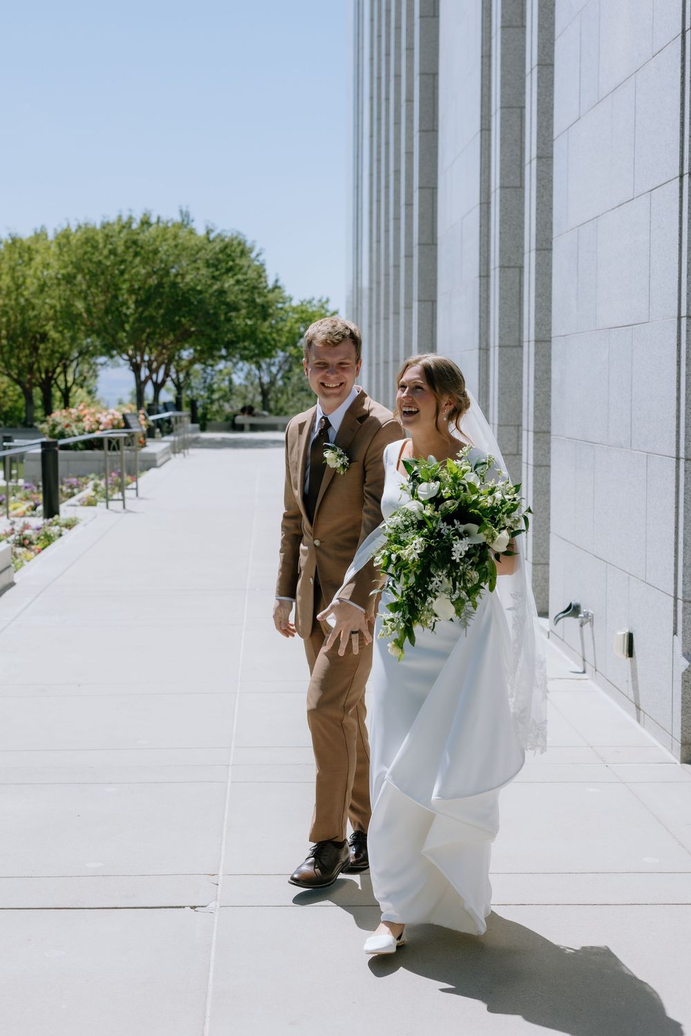 Happy bride and groom walking outside a wedding venue, bride holding a lush green and white bouquet on a sunny day.