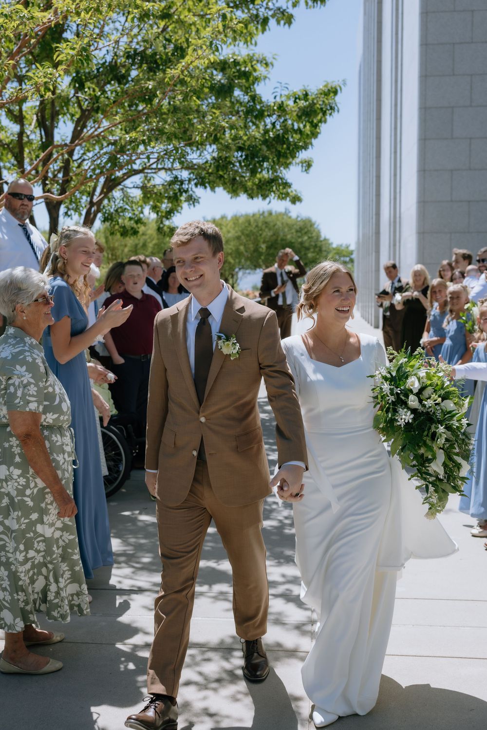 Smiling bride and groom walk down the aisle in sunlight as guests applaud their outdoor wedding ceremony.