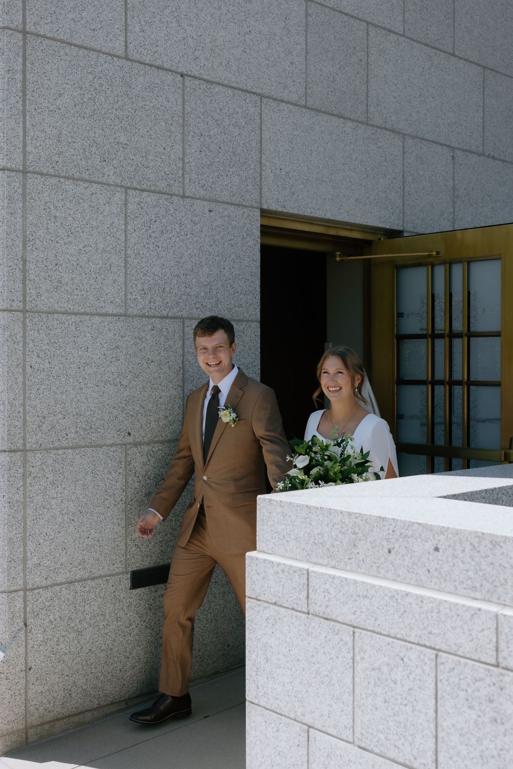 Smiling bride and groom in brown suit exit through golden doors of an LDS temple on their wedding day.