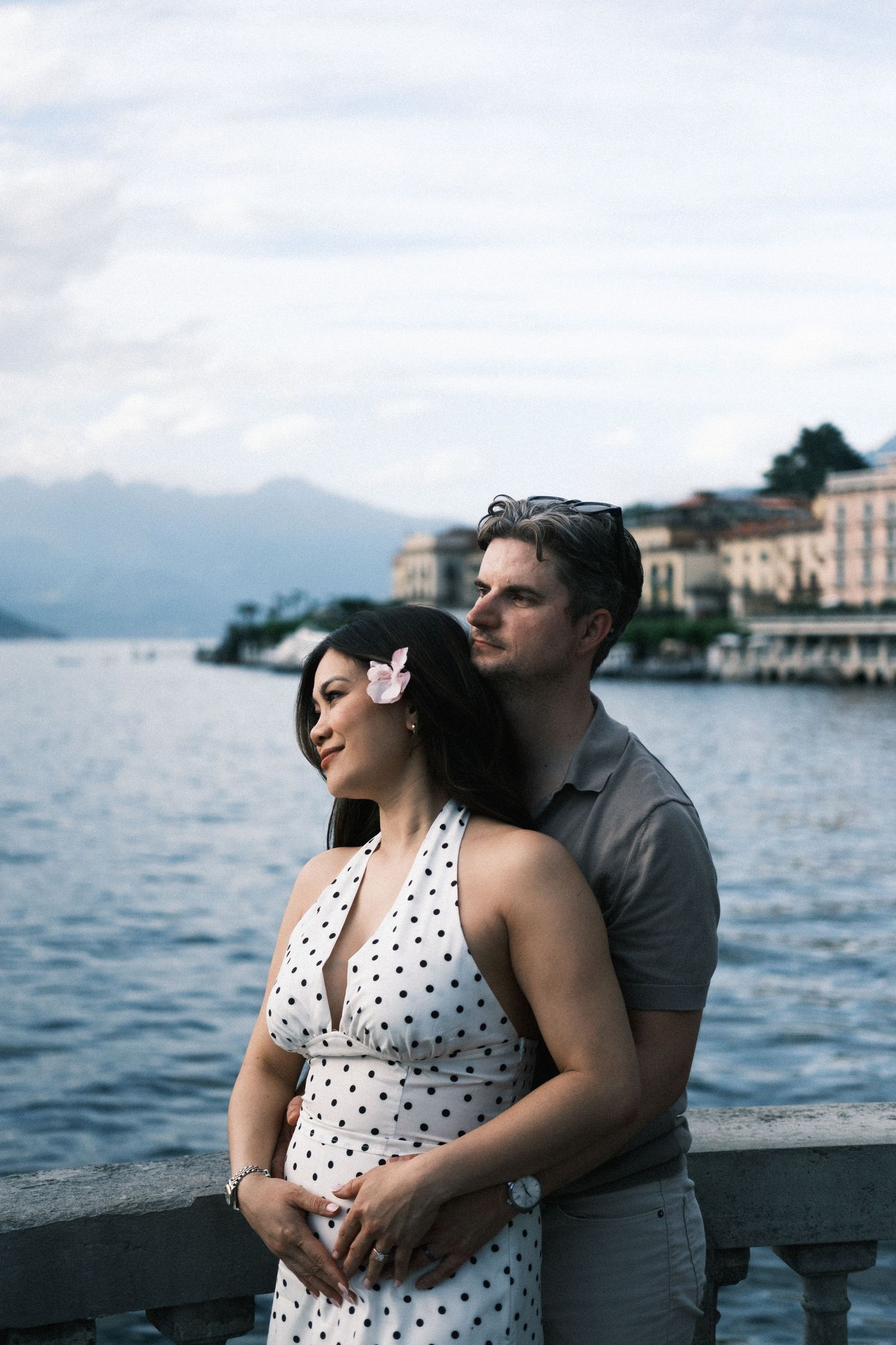 Couple embracing on a lakeside promenade with mountains and elegant buildings in the background.