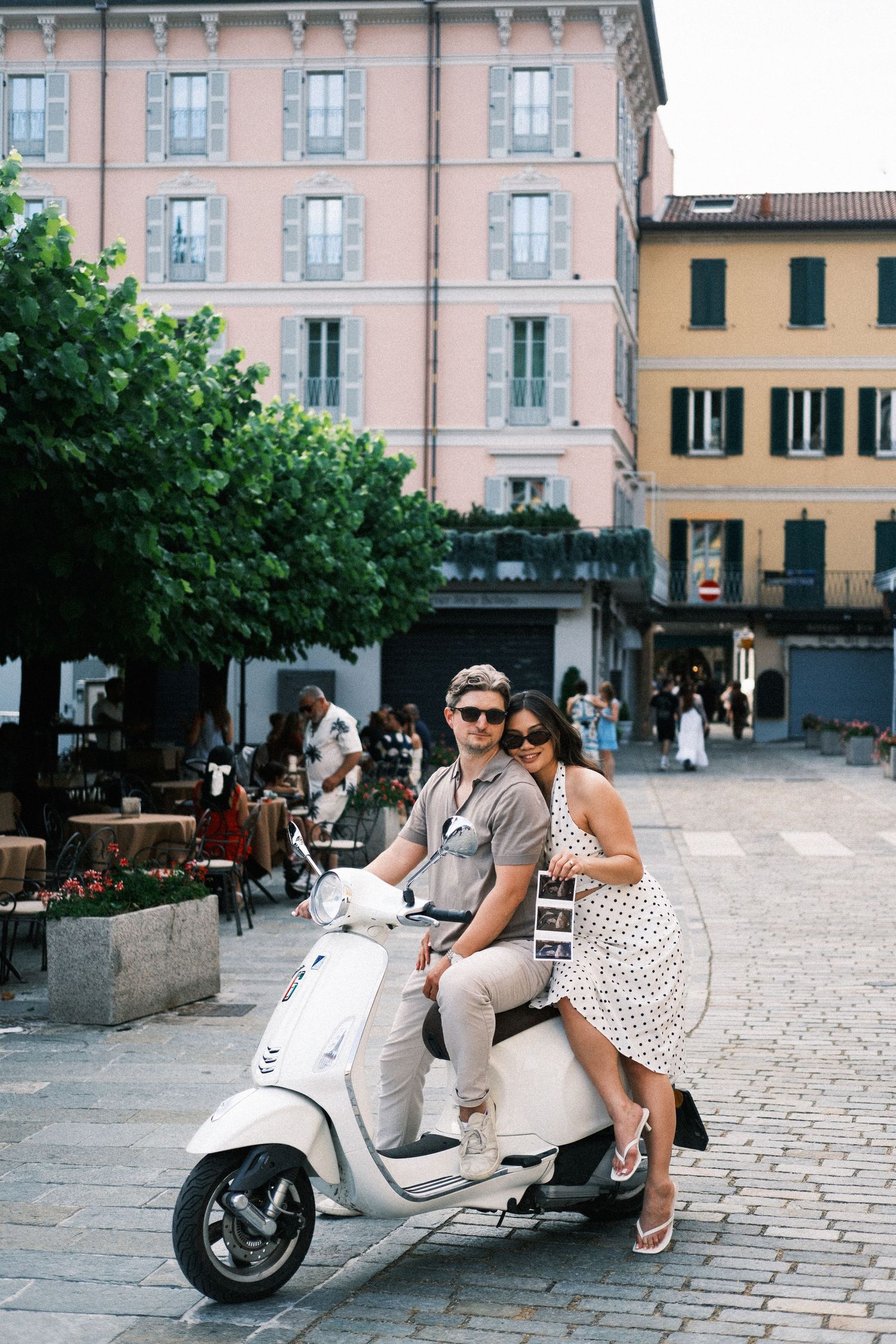 Couple riding a white Vespa scooter in a charming Italian piazza surrounded by colorful buildings and outdoor cafes.