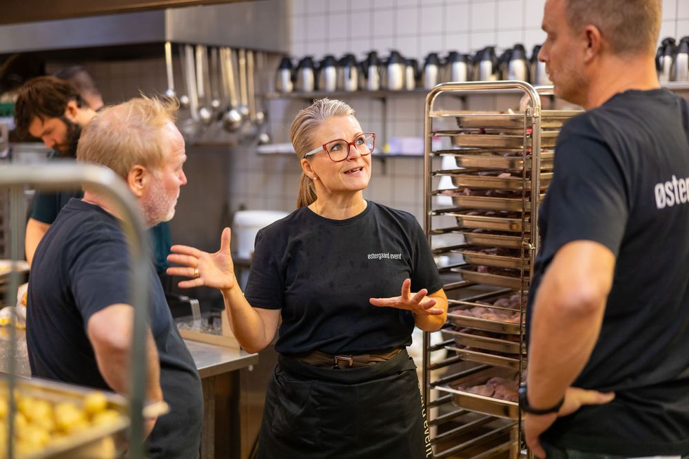 Kitchen staff in black uniforms discussing food preparation near a rack of baking trays in a professional kitchen.